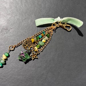 May include: A gold-colored keychain with multiple charms. The charms include a tree of life, a star, and a green and yellow gemstone cluster. A green ribbon is attached to the top of the keychain.