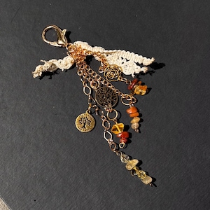 May include: A gold-colored keychain with a lobster clasp, adorned with a cream-colored lace ribbon and multiple charms. The charms include a tree of life pendant, a rose, and various amber-colored gemstone beads. The keychain is designed with a bohemian aesthetic.