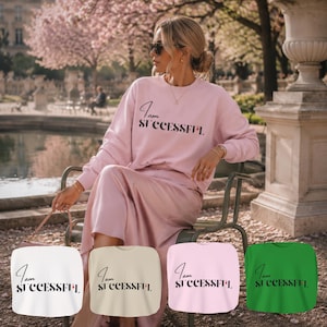 Könnte beinhalten: Eine Frau trägt ein rosa Sweatshirt und einen Rock mit dem Text "I am SUCCESSFUL" in schwarzer Schrift. Das Bild zeigt auch vier Sweatshirts in verschiedenen Farben: Weiß, Beige, Rosa und Grün, jeweils mit dem gleichen Text.
