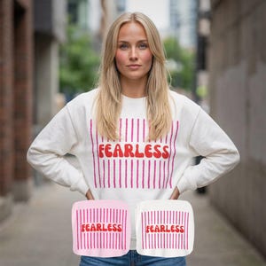 Könnte beinhalten: Ein weißes Sweatshirt mit dem Wort "FEARLESS" in Rot, umrahmt von vertikalen rosa Linien. Das Bild zeigt auch zwei kleine quadratische Designs mit demselben Text und Design, eines rosa und eines weiß.