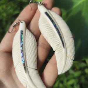 Op de afbeelding: Crèmekleurige veer oorbellen met iriserende abalone schelpinleg. De oorbellen hebben een gedetailleerd veerontwerp met gesneden lijnen en zilverkleurige oorhaaksluitingen. Een uniek accessoire.