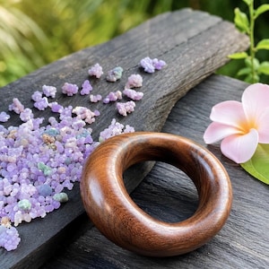 May include: A smooth, wooden ring with a rich, brown finish. The ring sits on a dark, textured surface, next to small, purple-colored crystals and a pink flower with a yellow center. The background is blurred with green foliage.
