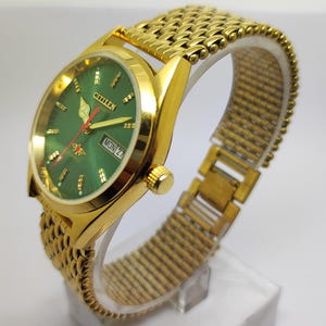 May include: A gold-tone Citizen automatic wristwatch featuring a round green dial with gold-tone hour markers, gold-tone hands, a red second hand, and a day-date window at the three o'clock position. The watch has a textured gold-tone metal link band.