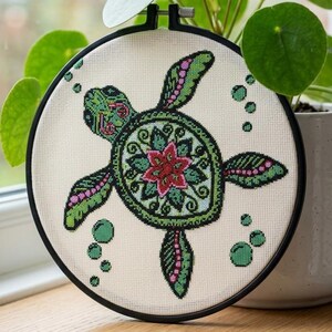 May include: A circular embroidery hoop containing a cross-stitch design of a sea turtle on cream-colored fabric. The turtle features a green shell with a central red floral pattern, green and pink flippers, and small green bubbles surrounding it.