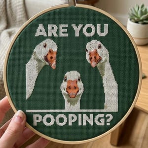 Peut inclure: Un tambour à broder circulaire en bois contenant un tissu vert foncé avec un motif au point de croix représentant trois oies blanches aux becs orange. Le texte "ARE YOU POOPING?" est brodé en lettres capitales blanches.