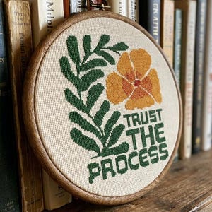 Peut inclure: Un tambour à broder circulaire en bois contenant un tissu couleur crème avec un motif au point de croix. Le motif représente une feuille verte semblable à une fougère, une fleur orange et le texte "TRUST THE PROCESS" en lettres vertes.