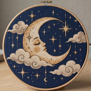 May include: A circular wooden embroidery hoop containing a cross-stitch design on dark blue fabric. The design features a golden crescent moon with a sleeping face, surrounded by beige clouds and various golden stars against the dark background.