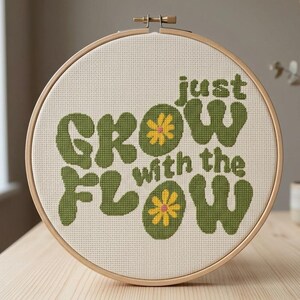 May include: A circular wooden embroidery hoop containing a piece of cream-colored Aida fabric. The fabric features a cross-stitch design with the green text "just grow with the flow" and two small yellow flowers with pink centers.