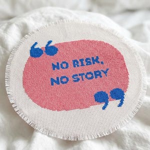 Könnte beinhalten: Ein kreisförmiges Kreuzstich-Stück auf weißem Stoff mit ausgefranstem Rand. Es zeigt eine rosa Sprechblasenform mit dem blauen Text 'NO RISK, NO STORY' und blauen Anführungszeichen in der oberen linken und unteren rechten Ecke der Sprechblase.