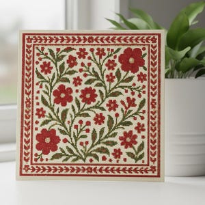 May include: A framed cross-stitch floral design. The artwork features a cream-coloured background with a pattern of red flowers and green leaves. The border is a deep red with a repeating leaf pattern. The piece is displayed near a window and a potted plant.