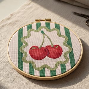 Pode incluir: Um bastidor de bordado circular de madeira contendo um design em ponto cruz de duas cerejas vermelho-brilhantes com caules verdes. O fundo apresenta listras verticais rosa-claro e verde-escuro, com uma borda verde recortada ao redor.