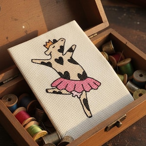 以下が含まれることがあります： ピンクのチュチュと金の王冠をつけた牛のクロスステッチ刺繍。木製の箱の中にあります。牛には黒い斑点とハート型の模様があります。箱には糸巻きと指ぬきが入っており、クラフトテーマを示唆しています。