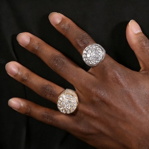 Puede incluir: Dos anillos con un grupo de pequeñas piedras transparentes. Uno es dorado y el otro plateado. Los anillos están en una mano de piel oscura, con un fondo negro.
