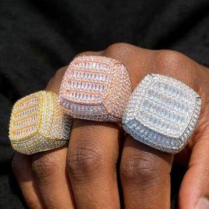 May include: Three square-faced rings, each encrusted with small, sparkling stones. One ring is gold-toned, another is rose gold, and the third is silver-toned. The rings are displayed on a dark background.