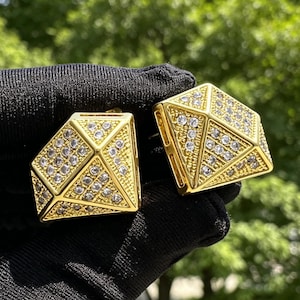 Puede incluir: Un par de pendientes dorados con forma de diamantes facetados. Cada pendiente presenta una estructura geométrica piramidal con una superficie dorada texturizada incrustada con numerosas piedras pequeñas, redondas y transparentes.
