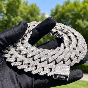 May include: A silver-toned cuban link chain necklace featuring a thick, interlocking design completely encrusted with clear, sparkling stones. The necklace includes a rectangular box clasp closure that is also covered in matching stones.