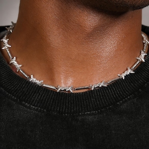 Op de afbeelding: Een zilverkleurige ketting met een prikkeldraadontwerp. De ketting is gemaakt van kleine, sprankelende stenen en wordt om de nek gedragen. De achtergrond is een donkere, effen kleur.