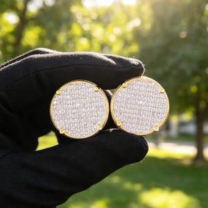 May include: A pair of circular gold-tone earrings featuring a pavé setting of numerous small, clear, reflective stones. Each earring has a textured, beaded border and four small prongs securing the stones within the round metal frame.