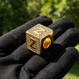 Può includere: Una perla cubica color oro che presenta la lettera Z su due lati visibili. Le superfici sono incastonate con piccole pietre chiare e scintillanti, e un foro circolare attraversa il centro della perla per l'inserimento su un cordino.