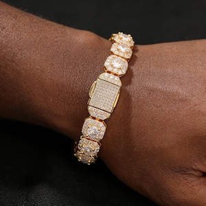 Puede incluir: Una pulsera dorada con un cierre rectangular y eslabones cuadrados. Cada eslabón presenta una piedra grande, transparente y de talla redonda, rodeada de piedras más pequeñas. El cierre está cubierto de pequeñas piedras brillantes.