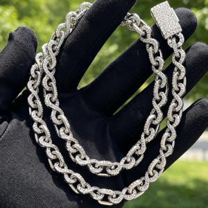 Puede incluir: Un collar de cadena en tono plateado con eslabones entrelazados en forma de infinito incrustados con pequeñas piedras transparentes. El collar incluye un cierre de caja rectangular también cubierto de pequeñas piedras transparentes, creando una apariencia uniforme y brillante en toda la pieza.
