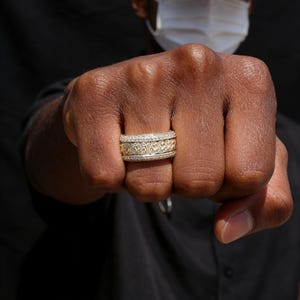 May include: A gold and diamond ring with a textured design. The ring is shown on a person's fist, with a black background. The ring has a band of diamonds and a textured gold center.