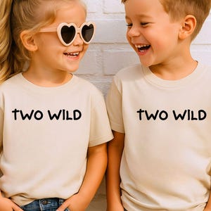 May include: Two children wearing matching beige t-shirts with the words "TWO WILD" in black. The girl is wearing heart-shaped sunglasses and a bow in her hair. Both children are smiling and wearing denim shorts.