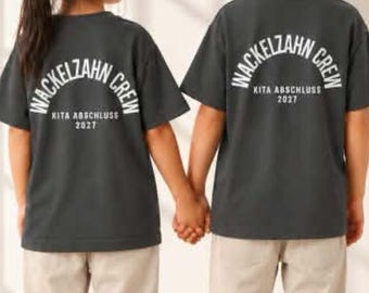 Puede incluir: Dos camisetas de manga corta de color gris oscuro con texto blanco en la espalda que dice WACKELZAHN CREW KITA ABSCHLUSS 2027. Las camisetas se combinan con pantalones de color beige claro y zapatillas blancas, vistas desde atrás.