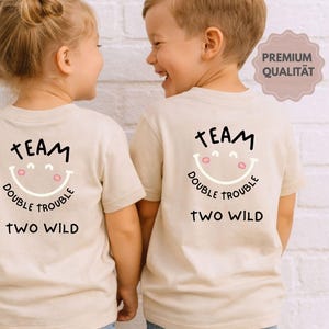 May include: Two children wearing matching beige t-shirts with the text "TEAM DOUBLE TROUBLE TWO WILD" and a smiling face graphic. The shirts are short-sleeved and the children are wearing blue jeans.