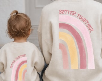 Puede incluir: Dos sudaderas beige de cuello redondo con un gráfico en la espalda que muestra un arcoíris en tonos rosa, amarillo y malva. El texto "BETTER TOGETHER" está impreso en una fuente curva de color rosa oscuro sobre el arcoíris.