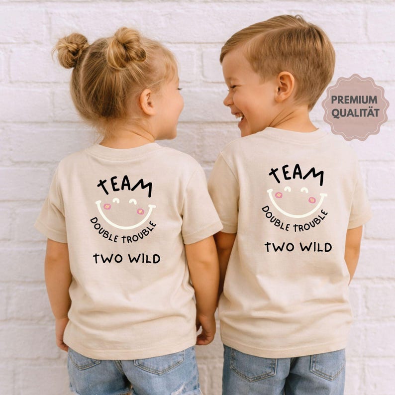 May include: Two children wearing matching beige t-shirts with the text "TEAM DOUBLE TROUBLE TWO WILD" and a smiling face graphic. The shirts are short-sleeved and the children are wearing blue jeans.