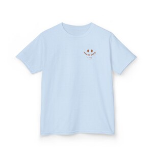 Puede incluir: Una camiseta de manga corta de color azul claro con un peque&ntilde;o gr&aacute;fico marr&oacute;n en la parte superior izquierda del pecho. El gr&aacute;fico consta de dos ojos ovalados sobre el texto "SCHULKIND" en una fuente curva, con el nombre "Finn" debajo.