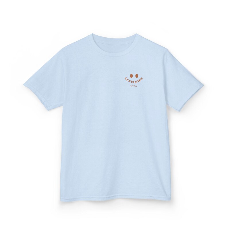 Puede incluir: Una camiseta de manga corta de color azul claro con un peque&ntilde;o gr&aacute;fico marr&oacute;n en la parte superior izquierda del pecho. El gr&aacute;fico consta de dos ojos ovalados sobre el texto "SCHULKIND" en una fuente curva, con el nombre "Finn" debajo.