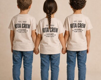 Puede incluir: Tres camisetas de color beige con texto negro en la espalda que dice EST. 2021 KITA CREW FRIENDS FOR LIFE, combinadas con vaqueros azules y zapatillas blancas. Las camisetas tienen cuello redondo y manga corta, usadas por tres personas.