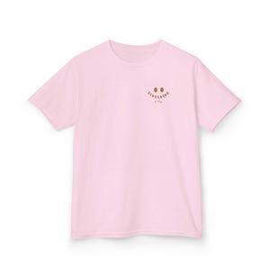 Puede incluir: Una camiseta de manga corta de color rosa claro con un peque&ntilde;o gr&aacute;fico marr&oacute;n en la parte superior izquierda del pecho. El gr&aacute;fico consta de dos ojos ovalados marrones sobre el texto "SCHULKIND" en una fuente curva marr&oacute;n, con el nombre "Finn" debajo.