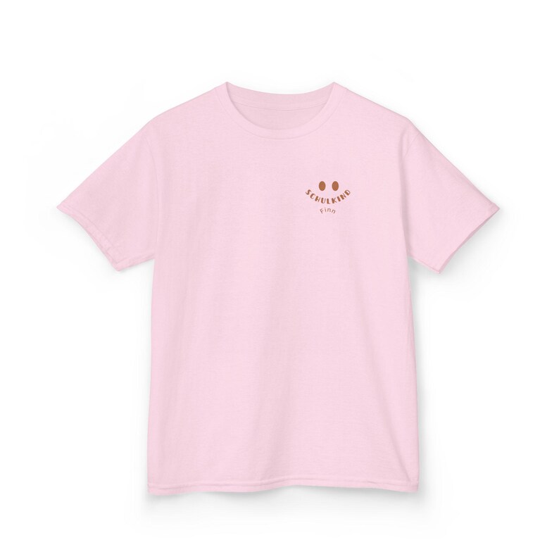 Puede incluir: Una camiseta de manga corta de color rosa claro con un peque&ntilde;o gr&aacute;fico marr&oacute;n en la parte superior izquierda del pecho. El gr&aacute;fico consta de dos ojos ovalados marrones sobre el texto "SCHULKIND" en una fuente curva marr&oacute;n, con el nombre "Finn" debajo.