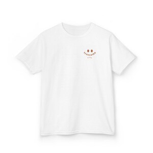 Puede incluir: Una camiseta blanca de manga corta con un peque&ntilde;o gr&aacute;fico marr&oacute;n en la parte superior izquierda del pecho. El gr&aacute;fico consta de dos ojos ovalados sobre el texto SCHULKIND y el nombre Finn en una fuente curva, marr&oacute;n y sin remates.