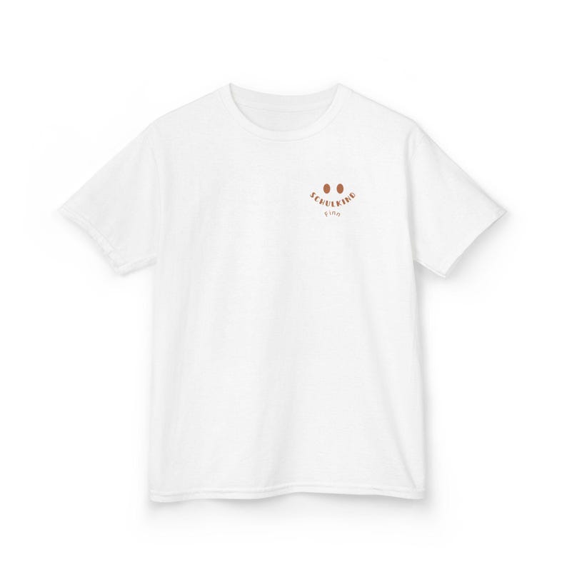 Puede incluir: Una camiseta blanca de manga corta con un peque&ntilde;o gr&aacute;fico marr&oacute;n en la parte superior izquierda del pecho. El gr&aacute;fico consta de dos ojos ovalados sobre el texto SCHULKIND y el nombre Finn en una fuente curva, marr&oacute;n y sin remates.