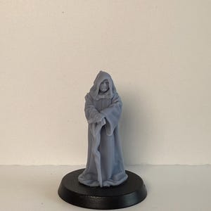 May include: A light grey figurine of a robed figure with a hood, holding a staff. The figure stands on a black circular base. The figurine is a miniature representation of a character from a science fiction franchise.