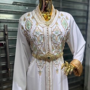 May include: A white caftan with gold and green embroidery, a gold belt with a green gem, and a pearl necklace. The caftan has long sleeves and is buttoned down the front. The mannequin's hands and head are gold.