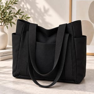 May include: A solid black tote bag featuring a structured rectangular silhouette, two long fabric shoulder straps, a large exterior slip pocket on the front, and two side pockets. The bag is constructed from a matte fabric with reinforced stitching.