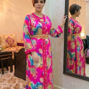 Peut inclure: Un caftan rose vif avec un motif floral coloré et des détails dorés. Le vêtement à manches longues est orné d'une ceinture et de bordures dorées, et est porté avec des bijoux dorés. Le caftan est un vêtement traditionnel, souvent porté pour les occasions spéciales.