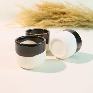 Könnte beinhalten: Drei kleine Keramikbecher, weiß mit einem schwarzen Band am oberen Rand. Einer der Becher liegt auf der Seite. Die Becher sind auf einer weißen Oberfläche angeordnet. Geeignet für Espresso oder kleine Getränke.