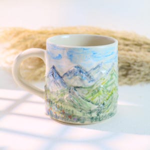 Könnte beinhalten: Weißer Keramikbecher mit handgemaltem Berglandschaftsdesign. Der Becher zeigt einen hellblauen Himmel mit flauschigen Wolken und grünen und blauen Bergen. Der Griff ist weiß und der Becher ist etwa 10 cm hoch.