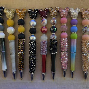 May include: A collection of nine pens featuring decorative toppers shaped like mouse ears. Each pen is adorned with various colorful beads, including solid, metallic, and rhinestone-encrusted spheres, arranged in unique patterns along the pen barrels.