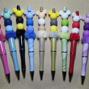 May include: A collection of ten pens arranged in a row, each featuring a mouse-ear topper, two round beads, and a fluffy pom-pom accent. The pens display various colors including pink, blue, black, yellow, white, green, red, and purple patterns.