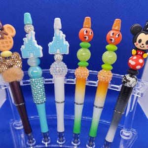 May include: A collection of decorative pens with various character-themed toppers. The pens feature designs such as a Mickey Mouse, a castle, and pumpkin-shaped figures. The pens have colorful beads and glitter accents, with a clear plastic stand.
