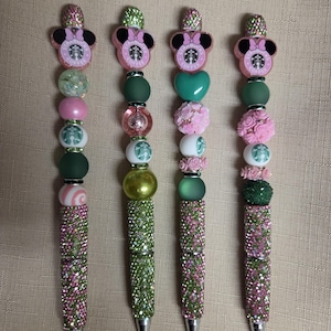 May include: Four pens featuring decorative beads in shades of pink and green. Each pen is topped with a pink Minnie Mouse-shaped charm featuring a Starbucks logo. The barrels are covered in pink and green rhinestones, with various round, heart-shaped, and floral-textured beads arranged along the upper portion of each pen.