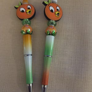 May include: Two decorative pens featuring orange bird toppers with green leaves and a white or orange top. The pen barrels have a gradient design, transitioning from green to orange. The pens have silver accents.