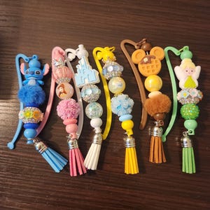 May include: A collection of beaded bookmarks with various charms, including a blue Stitch character, a pink rose, a castle, a Mickey Mouse head, and a fairy. Each bookmark has a tassel in a different color.